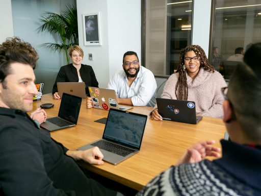 four people all on laptops, two men and two women, listen to person talking in a board meeting