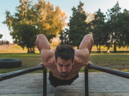 Shirtless man showcasing fitness by doing push-ups on metal bars in a park setting.