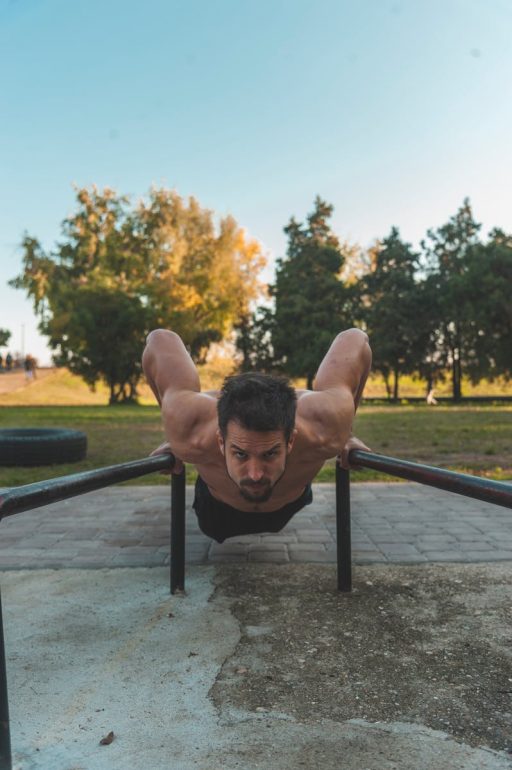 Shirtless man showcasing fitness by doing push-ups on metal bars in a park setting.