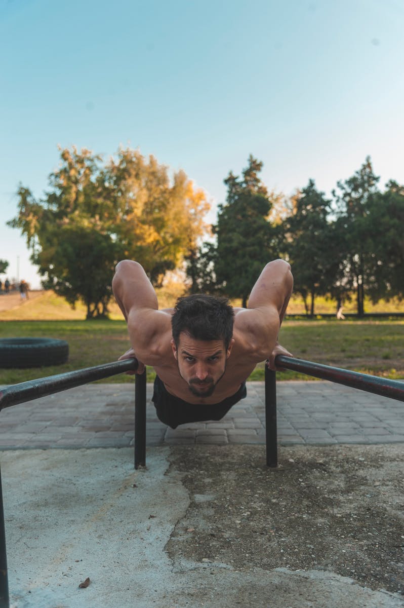 Shirtless man showcasing fitness by doing push-ups on metal bars in a park setting.